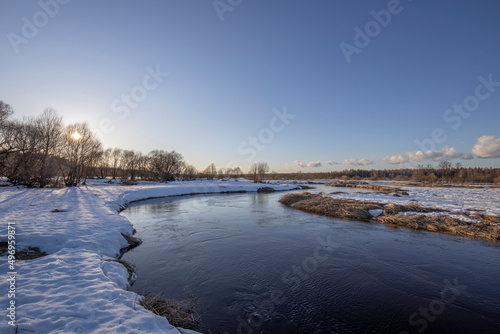A picturesque landscape, early spring, a river with snow-covered banks, dry grass and bushes. March sunny day by the river. The first thaws, the snow is melting. © Sergei