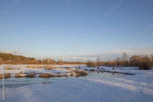 March sunny evening by the river. Blue sky over the horizon. A picturesque landscape, early spring, a river with snow-covered banks, dry grass and bushes. The first thaws, the snow is melting