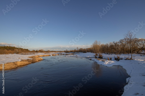 March sunny evening by the river. Blue sky over the horizon. A picturesque landscape  early spring  a river with snow-covered banks  dry grass and bushes. The first thaws  the snow is melting