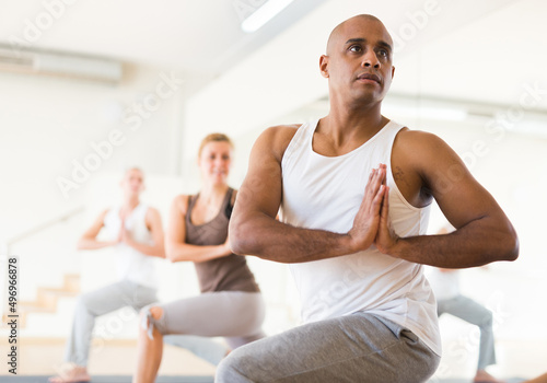 Concentrated Latino doing yoga with group of people in fitness studio, standing in twisting asana Parivritta Parsvakonasana