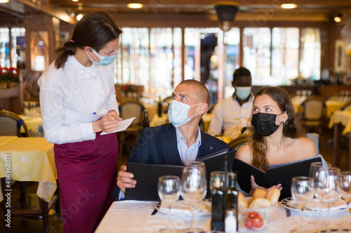 Waitress in face mask receiving order from couple of guests in masks choosing drinks and meals in restaurant