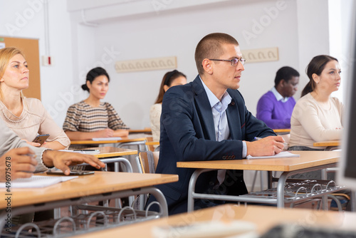 Intelligent adult man in glasses attentively listening and making notes of lecture in classroom full of students