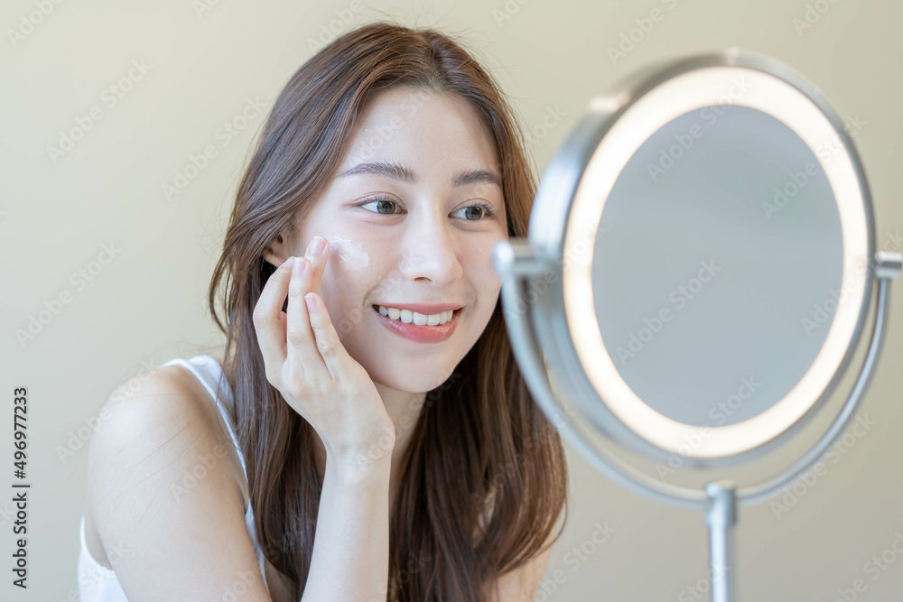 Fresh healthy skin, beautiful smile of asian young woman, girl looking at mirror, applying moisturizer on her face, putting cream treatment before make up cosmetic routine at home. Facial Beauty. Fresh healthy skin, beautiful smile of asian young woman, girl looking at mirror, applying moisturizer on her face, putting cream treatment before make up cosmetic routine at home. Facial Beauty.