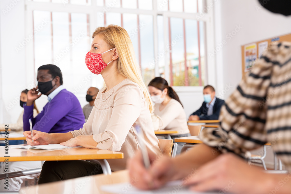Portrait of focused female in protective face mask sitting at desk studying in classroom with colleagues