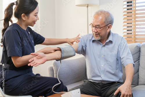 Pressure measurement of hypertension in elderly man sitting on couch, young woman nurse or caregiver care checking, measuring heart, blood by using sphygmomanometer monitor at home after retirement.