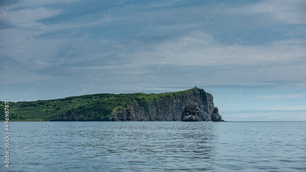 Kamchatka coast against a background of blue sky, clouds and ocean. Steep rocky slopes. Green vegetation on the hills. Ripples on the water. Avacha Bay