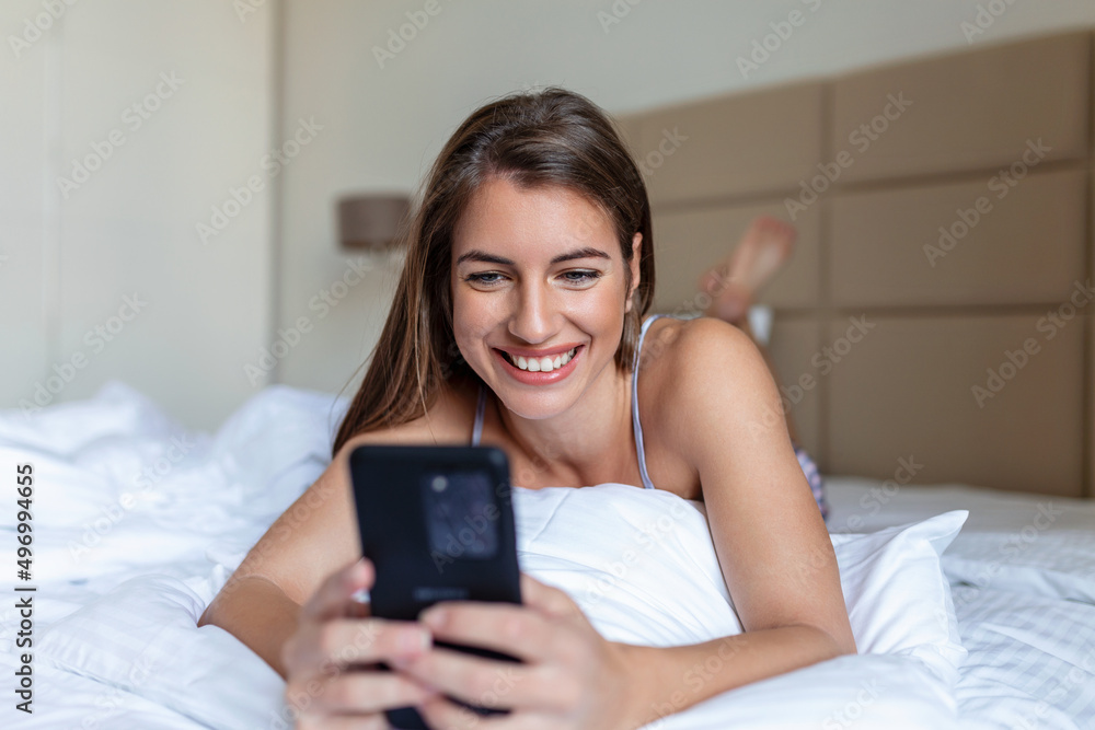 Smiling woman looking at mobile phone while laying on white bed. Happy brunette young woman using cellphone at home. Beautiful girl surfing the net on smartphone Smiling woman looking at mobile phone while laying on white bed. Happy brunette young woman using cellphone at home. Beautiful girl surfing the net on smartphone