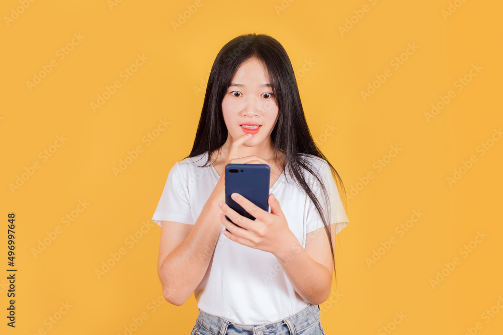 Shocked Asian woman wears white t-shirt using mobile phone on yellow background Shocked Asian woman wears white t-shirt using mobile phone on yellow background