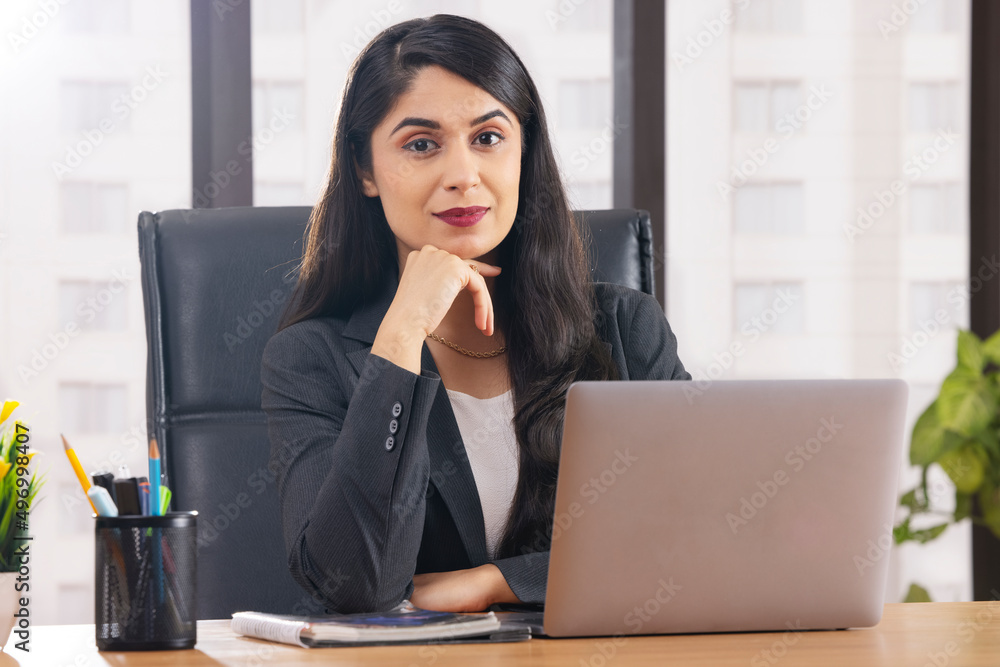 Portrait of a businesswoman looking at camera with hand on chin while working at desk in office using laptop Portrait of a businesswoman looking at camera with hand on chin while working at desk in office using laptop