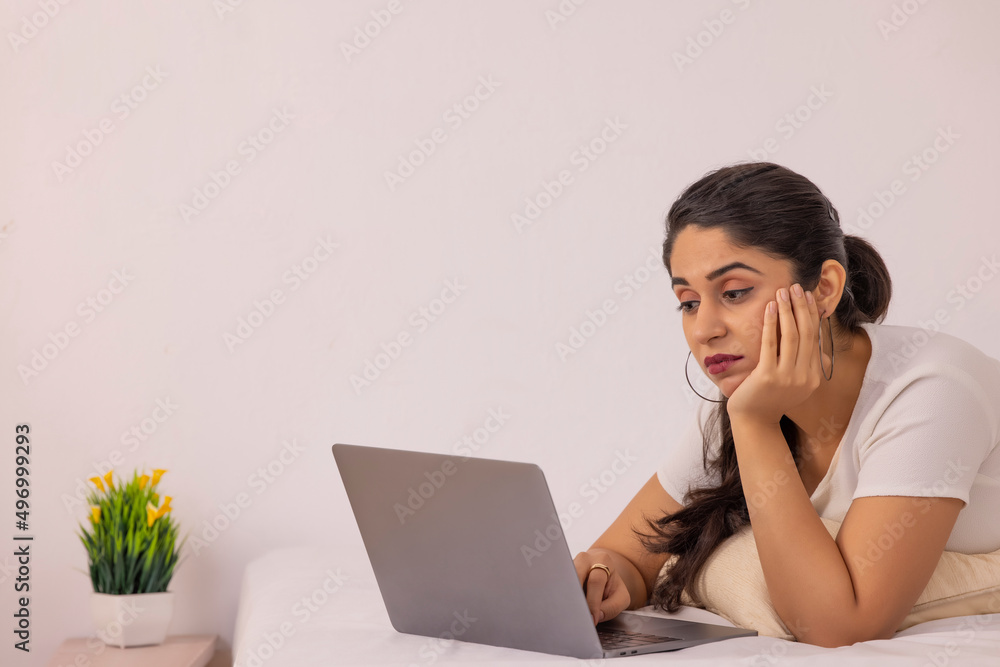 Portrait of young exhausted woman with hand on cheek using laptop while lying down on bed at home Portrait of young exhausted woman with hand on cheek using laptop while lying down on bed at home
