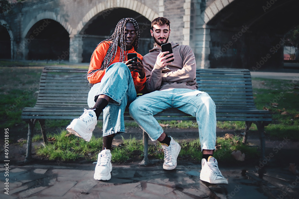 Obraz premium Couple fo young trendy biracial young people using smartphone sitting together on a bench in the public city park - young friends using social network and the internet - technology addiction concept
