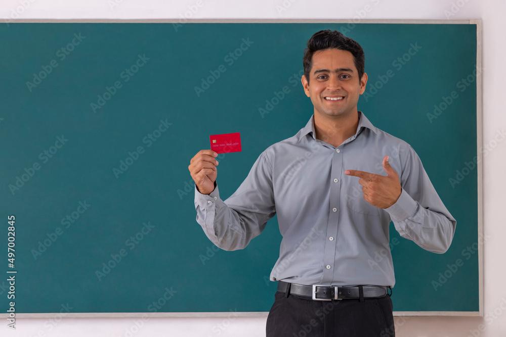 School teacher pointing at credit card while standing in front of blackboard in classroom School teacher pointing at credit card while standing in front of blackboard in classroom
