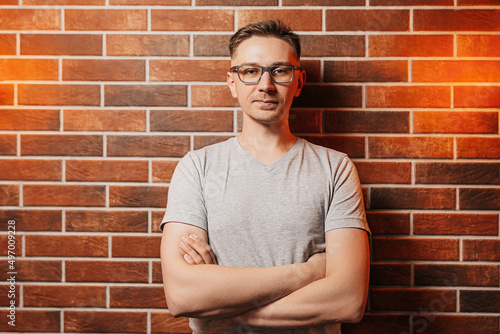 Handsome man smiling and posing against a red brick wall background. Lifestyle portrait of casual businessman with glasses. Professional guy smiles confident in gray t-shirt. Entrepreneurship concept