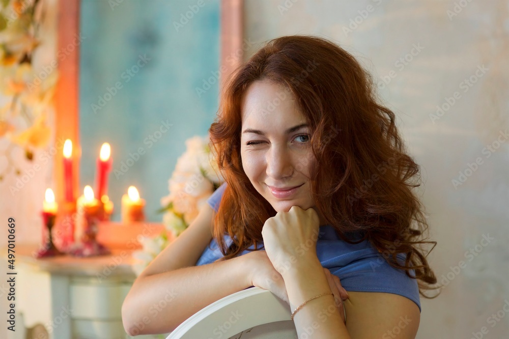 Naklejka premium A girl with red hair and a blue dress is sitting on a vintage chair indoors and winking with one eye.