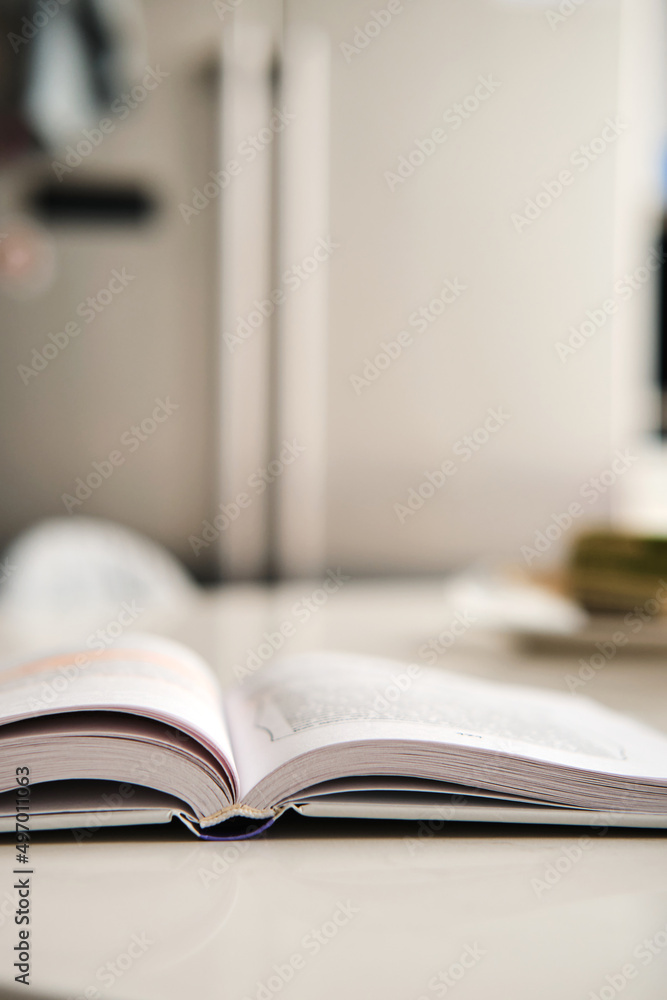 Open hardcover book with white pages lies on artificial stone table at kitchen, fridge on background. Books spine. Selective focus. Vertical stories background. Open hardcover book with white pages lies on artificial stone table at kitchen, fridge on background. Books spine. Selective focus. Vertical stories background.
