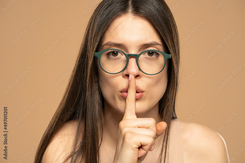 Portrait of young woman finger covering lips mouth do not tell look camera isolated on beige color background Portrait of young woman finger covering lips mouth do not tell look camera isolated on beige color background