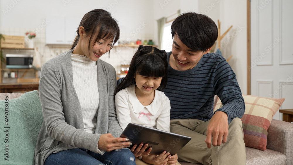 Cheerful Asian father, mother and little girl relaxing in living room with electric pad. The have good laugh while sitting together on sofa watching funny online show Cheerful Asian father, mother and little girl relaxing in living room with electric pad. The have good laugh while sitting together on sofa watching funny online show