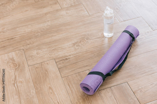 Rolled purple yoga mat and clear plastic water bottle on a wooden surface with natural light. Gender-neutral fitness yoga and exercise props at home or gym banner with copy space. Active lifestyle.