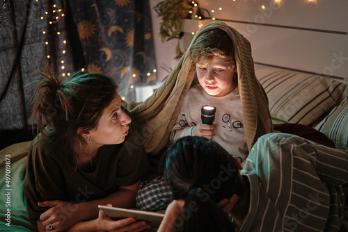 Fotografia Little boy sitting under blanket with flashlight and telling scary tales to his