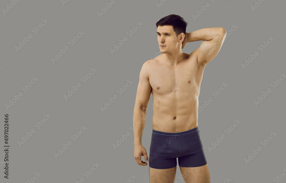 Handsome man with fit, slim body posing topless in underwear. Young male fashion model in comfortable underpants looking away standing with hand behind head isolated on gray copyspace background Handsome man with fit, slim body posing topless in underwear. Young male fashion model in comfortable underpants looking away standing with hand behind head isolated on gray copyspace background