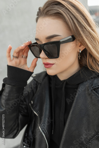 Urban fashionable portrait of young beautiful girl with stylish rock leather and black fashion hoodie wear a cool modern sunglasses and walks on the street