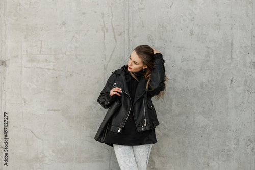 Beautiful young girl in fashionable clothes with a leather jacket, black hoodie, jeans, boots and a black leather bag stands near a gray concrete wall