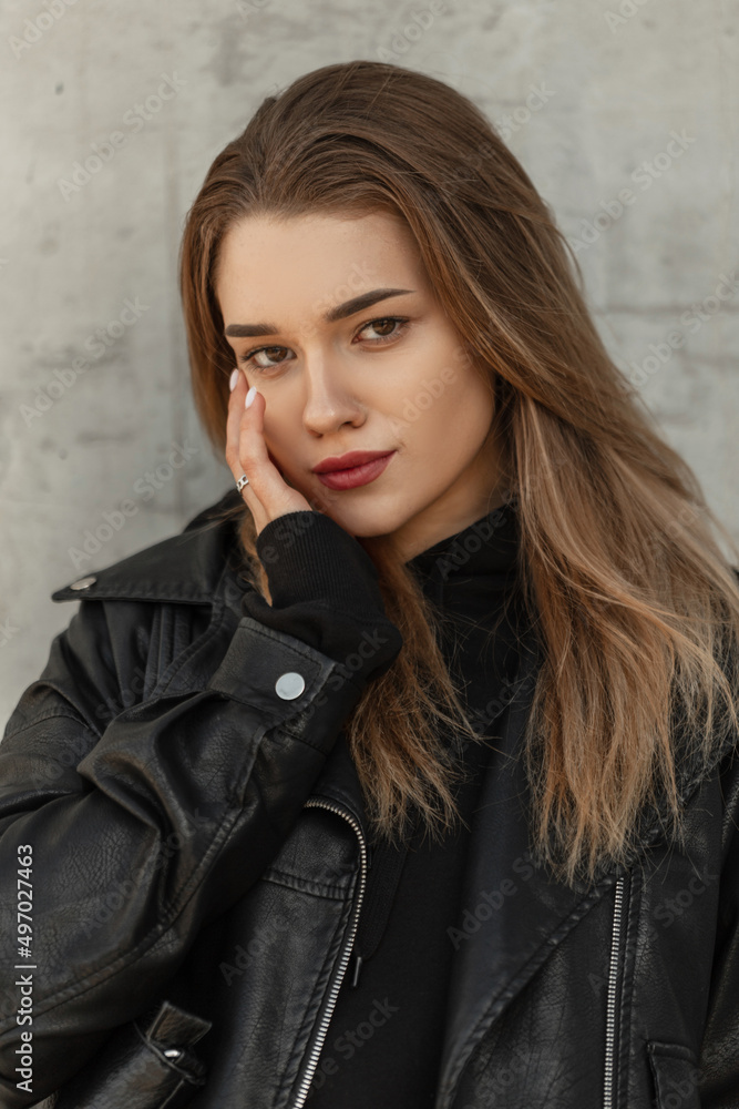Stylish beautiful young girl model hipster in fashionable clothes with a black leather jacket and a black hoodie on the street near a gray concrete vintage wall