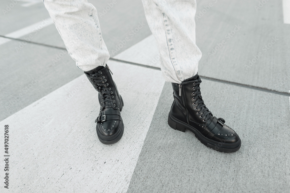 young stylish woman in fashionable black leather boots shoes with light-colored jeans is standing on the road on the street