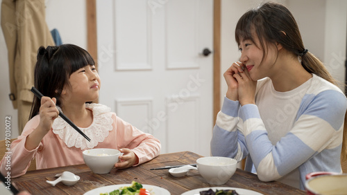 smiling asian mother looking her young daughter enjoying the meal she prepares at dining table at home. the girl saying yummy to her mom while eating