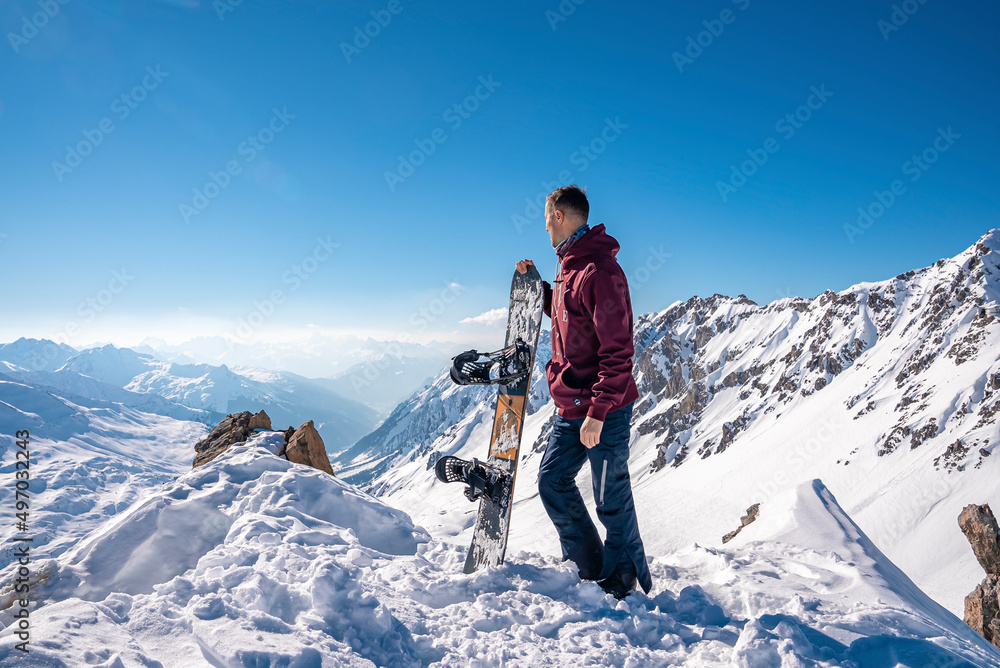 Young man holding snowboard while standing on top of snowy mountain on sunny day