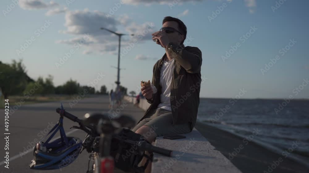 Cyclist sits on concrete fence near sea and drinks from disposable cup of coffee and eats sandwich on bicycle trip. Man has lunch after cycling near lake. Snack after work overlooking a large pond. Cyclist sits on concrete fence near sea and drinks from disposable cup of coffee and eats sandwich on bicycle trip. Man has lunch after cycling near lake. Snack after work overlooking a large pond.