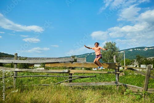 A school-age girl jumps from a wooden village fence surrounded by wild grasses and flowers. © Fotoproff
