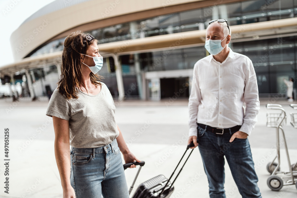 Travellers in protective masks chatting while leaving the airport with their luggage