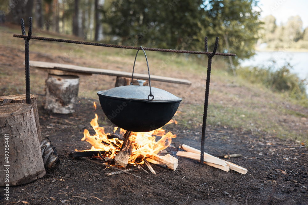 Cooking in cauldron on open fire in nature. Bowler on bonfire in forest. Pot from cast iron on campfire on background of river. Copy space. Cooking in cauldron on open fire in nature. Bowler on bonfire in forest. Pot from cast iron on campfire on background of river. Copy space.