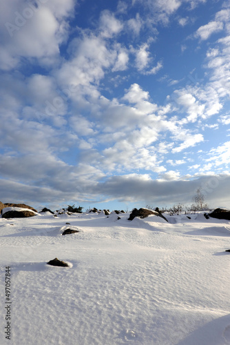 Winter tundra landscape with hilly snowy surface with stones and large boulders and long shadows from them under a beautiful blue sky with clouds © DyMaxFoto