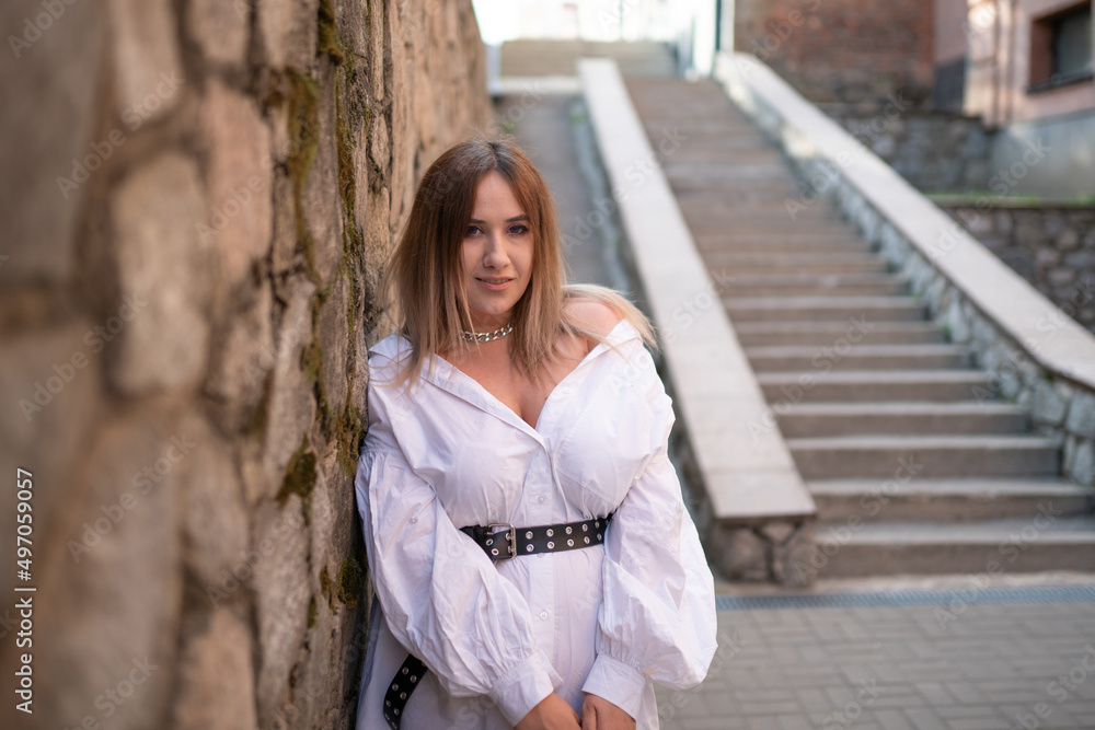 A very beautiful fashionable bohemian girl in a light white dress similar to a man's shirt, white sandals and a small handbag poses for a photographer in the city in summer A very beautiful fashionable bohemian girl in a light white dress similar to a man's shirt, white sandals and a small handbag poses for a photographer in the city in summer