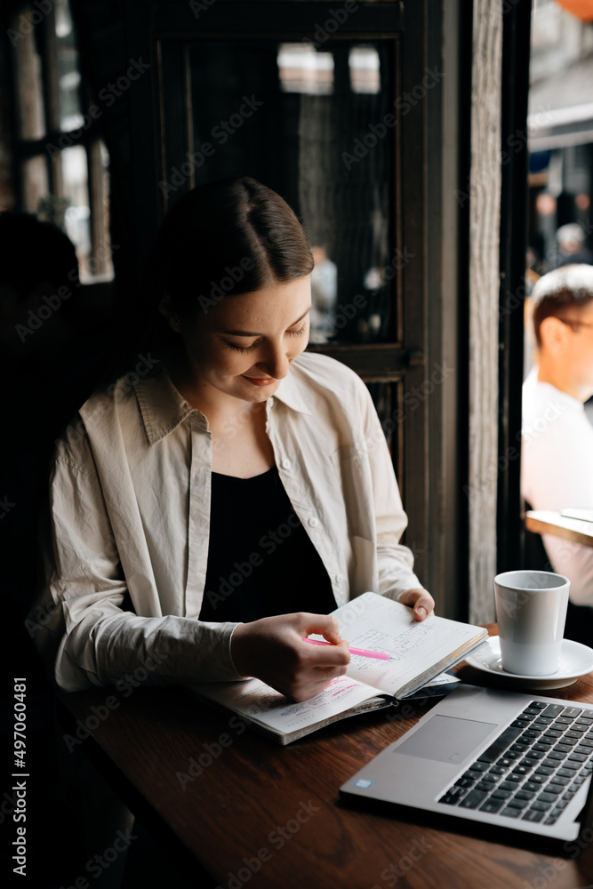 A young European freelancer girl with long dark hair in a shirt remotely works in a cafe, businesswoman are writing notes with pink pen, working on a laptop and drinking black coffee A young European freelancer girl with long dark hair in a shirt remotely works in a cafe, businesswoman are writing notes with pink pen, working on a laptop and drinking black coffee