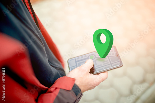 Online navigation, GPS and Geocaching. Green geolocation sign above the smartphone screen with an abstract online map. Close-up of a man's hand using a smartphone on the street photo