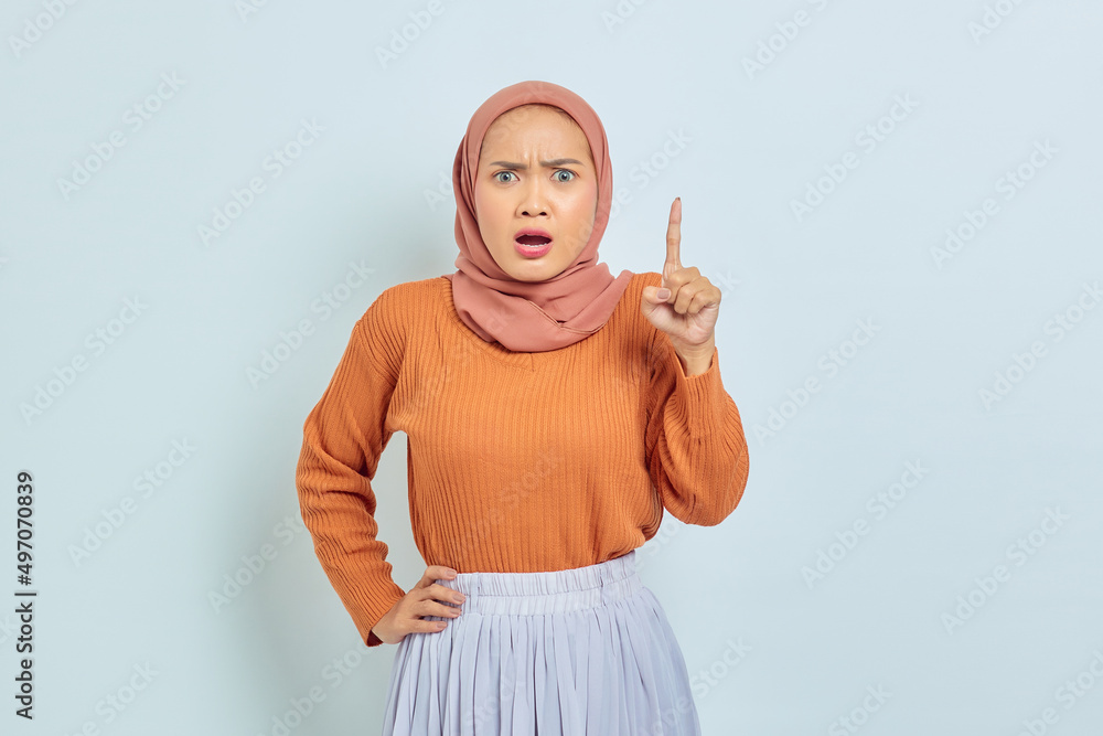 Naklejka premium Surprised young Asian Muslim woman in brown sweater pointing to copy space with finger isolated over white background. Muslim lifestyle concept