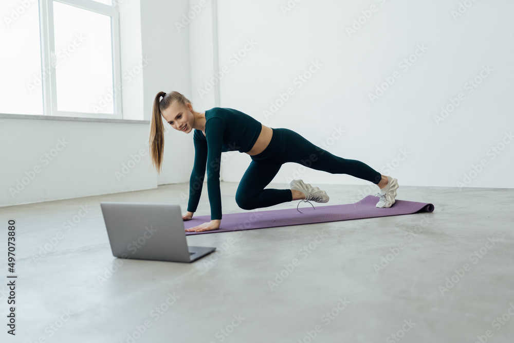 Fit woman doing yoga exercise while watching tutorial on laptop at home. Attractive woman exercising on a mat and watching instructional videos on laptop.