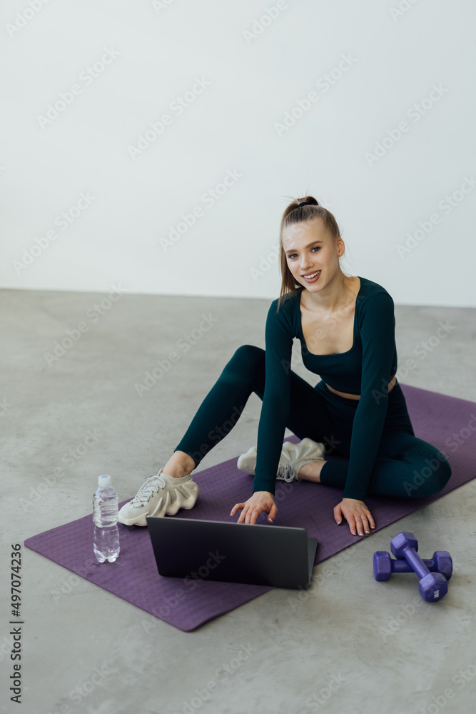 Fototapeta premium Training Concept. Smiling fit young woman in sports clothes watching fitness videos on internet using laptop near window, lying on floor yoga mat in the modern living room with minimal interior or gym