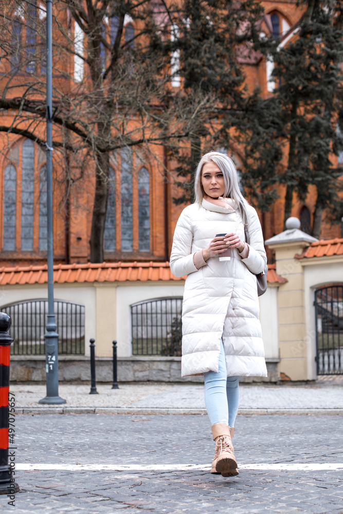 Portrait of young pretty serious woman with long grey hair holding takeaway cup of coffee, standing in white jacket.