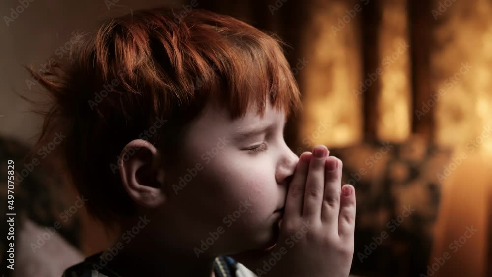 A young boy is praying in a dark room. A red-haired boy of seven to ten ...