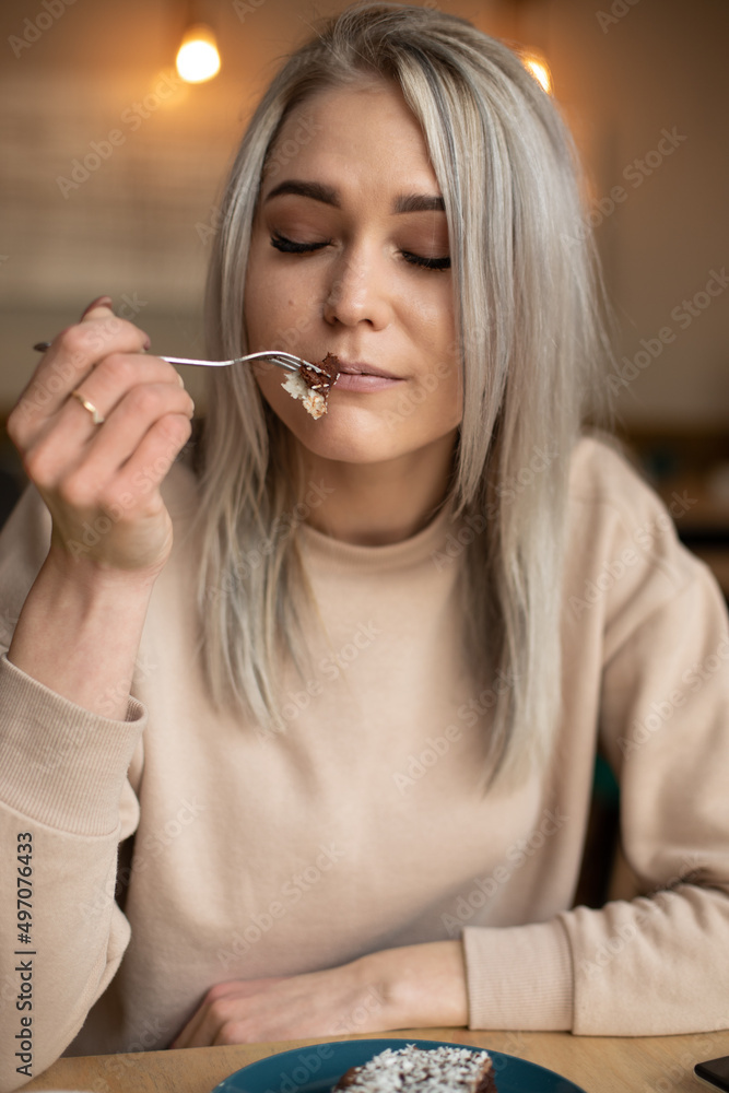 Portrait of young beautiful pleased woman with cute make-up holding fork with piece of cake, sitting at table in cafe. Portrait of young beautiful pleased woman with cute make-up holding fork with piece of cake, sitting at table in cafe.