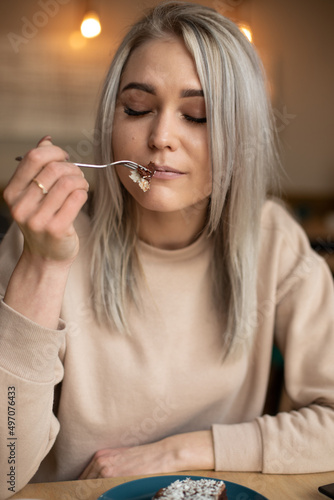 Portrait of young beautiful pleased woman with cute make-up holding fork with piece of cake, sitting at table in cafe. 