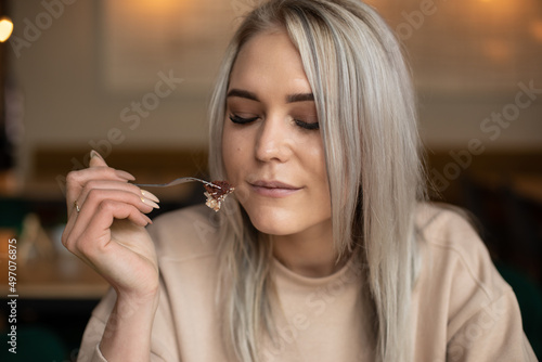 Portrait of young attractive pleased woman with long grey hair holding fork with piece of cake with closed eyes in cafe.