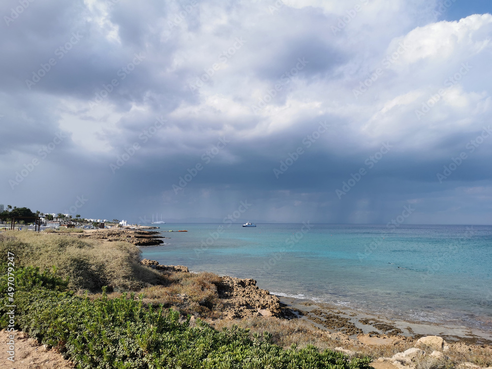 Sunny coast of the Mediterranean Sea with  stones, dramatic sky and rain above the sea, boats in the sea and the largest sailing yacht in the world.