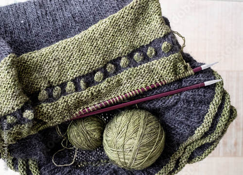 Green and black wool sweater in the process of being knitted. Photograph shows textured knitting including stripes and bobbles, knitting needles and balls of yarn.