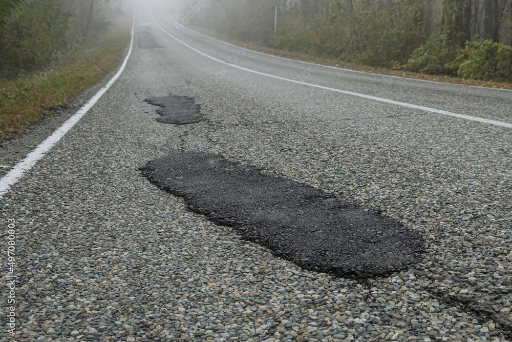 Naklejka premium Fresh patches after patching the road. Country highway in the foggy forest. Bad road condition.