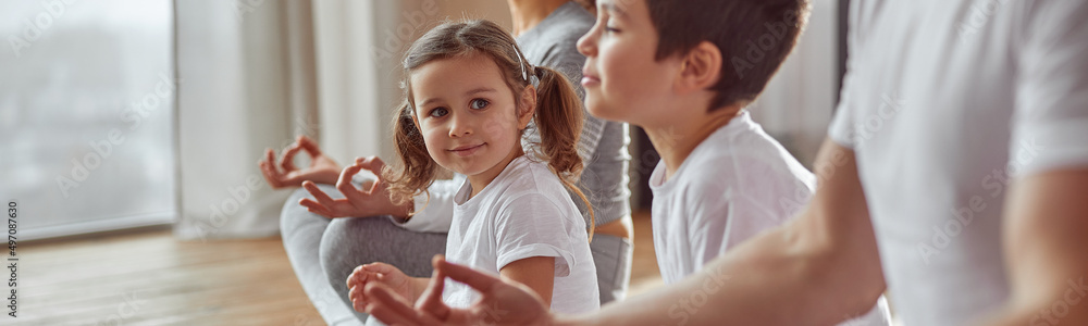 Cheerful daughter is sitting in lotus position near older brother and calm parents at home Cheerful daughter is sitting in lotus position near older brother and calm parents at home